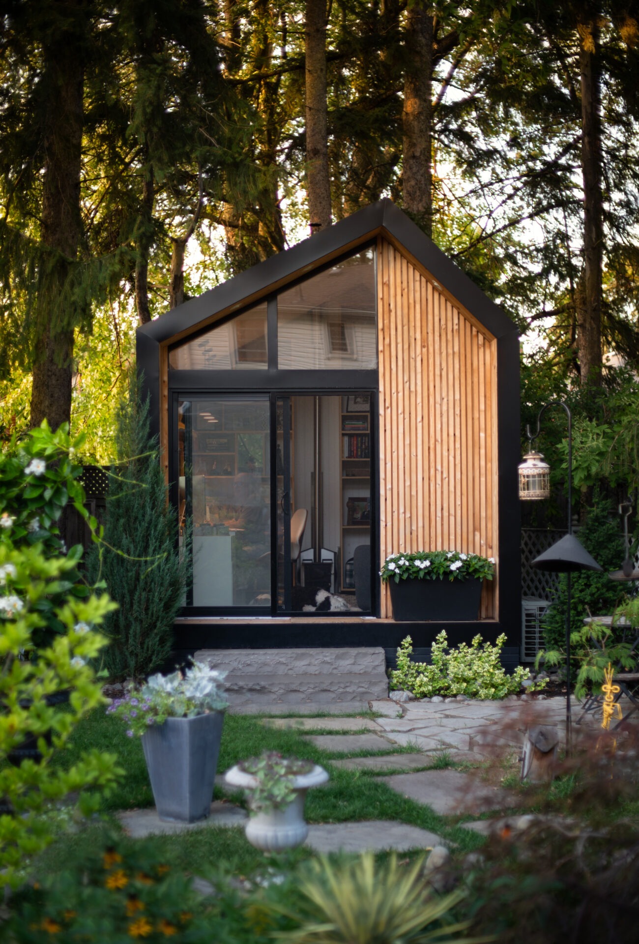 A modern garden cabin with wooden paneling, large glass doors, surrounded by lush greenery and a stone path leading to the entrance.