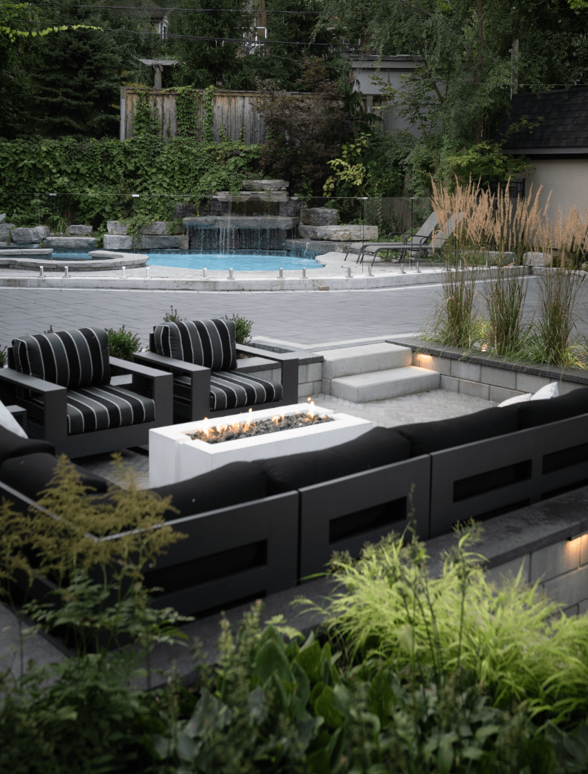 Modern outdoor patio with black furniture, fire pit, and garden. Adjacent is a waterfall pool surrounded by lush greenery. Peaceful, inviting setting.