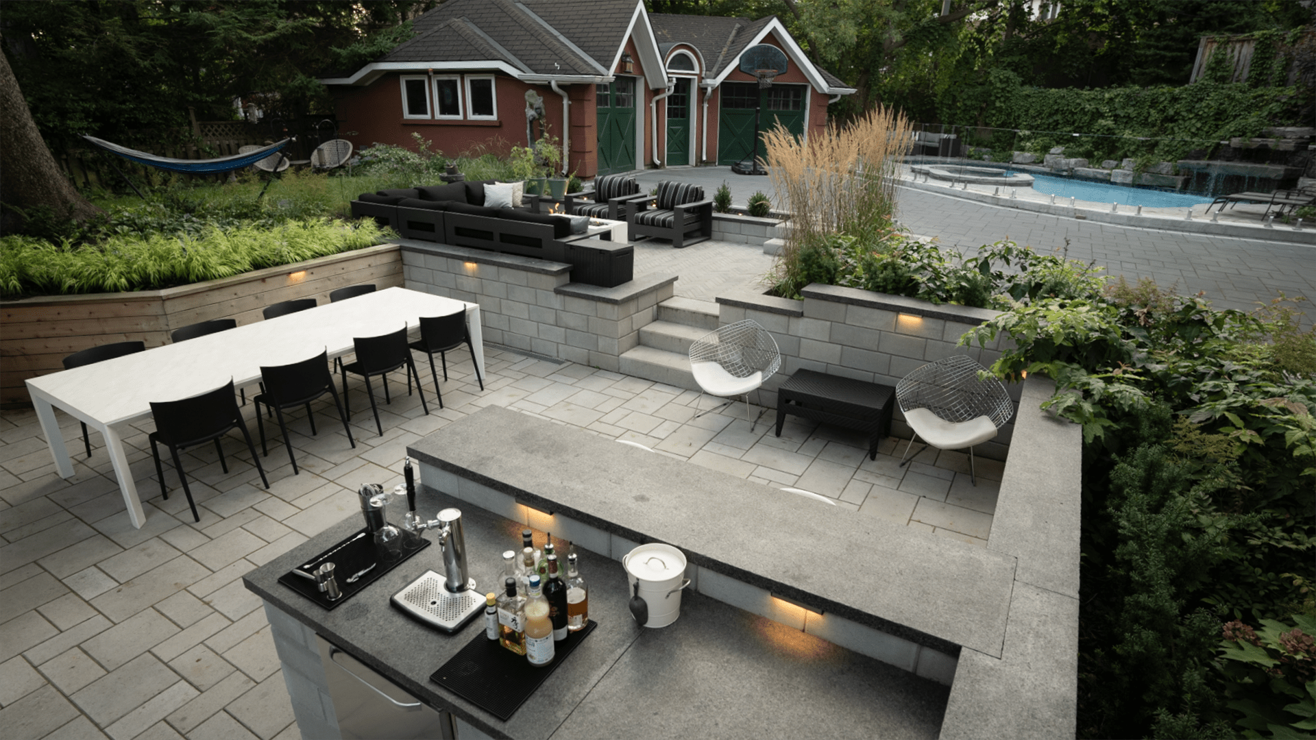 Modern backyard patio with a dining table, seating area, and outdoor kitchen. Features lush greenery and a pool near a red garden house.