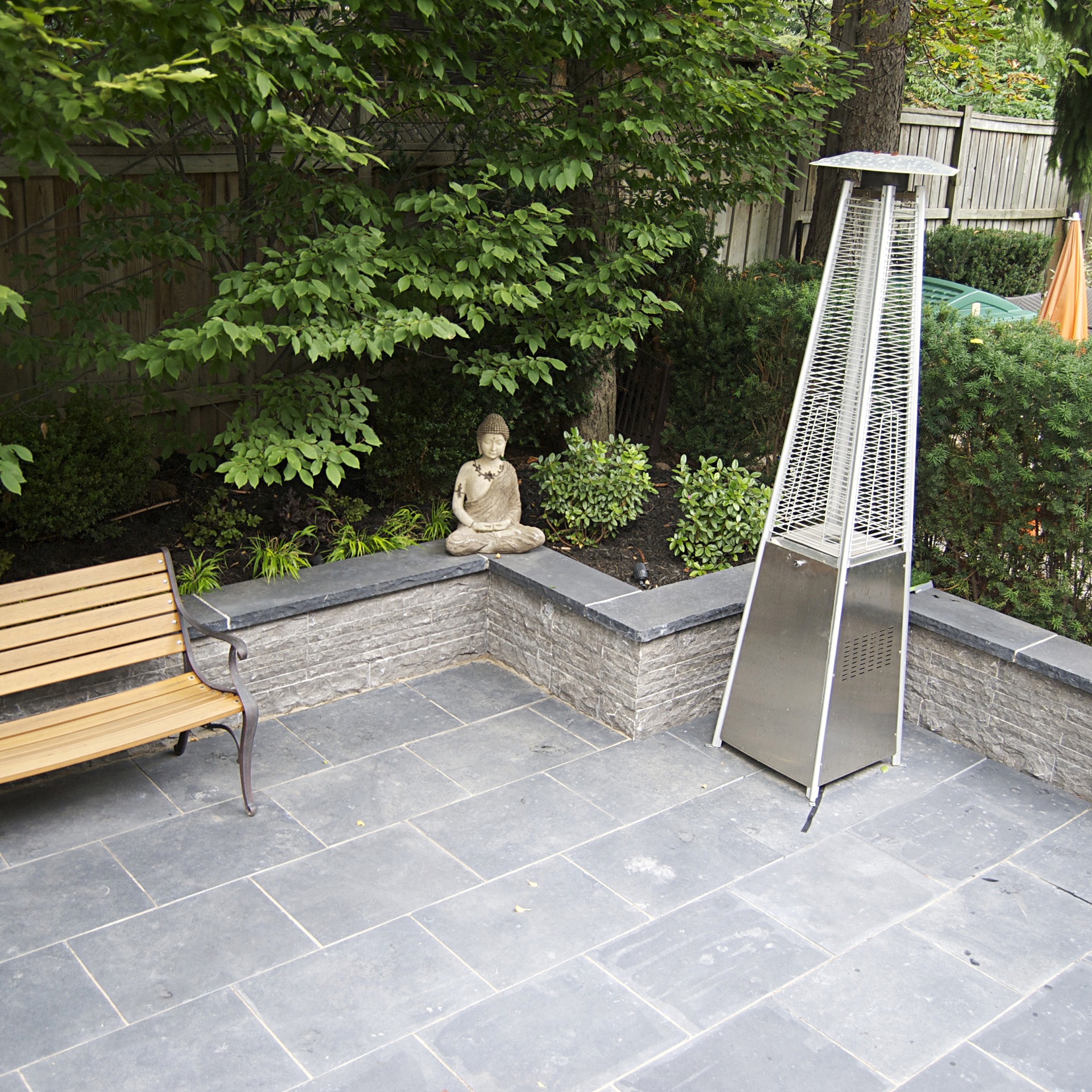 A serene patio features a wooden bench, Buddha statue, and outdoor heater, surrounded by lush greenery and stone tiles.