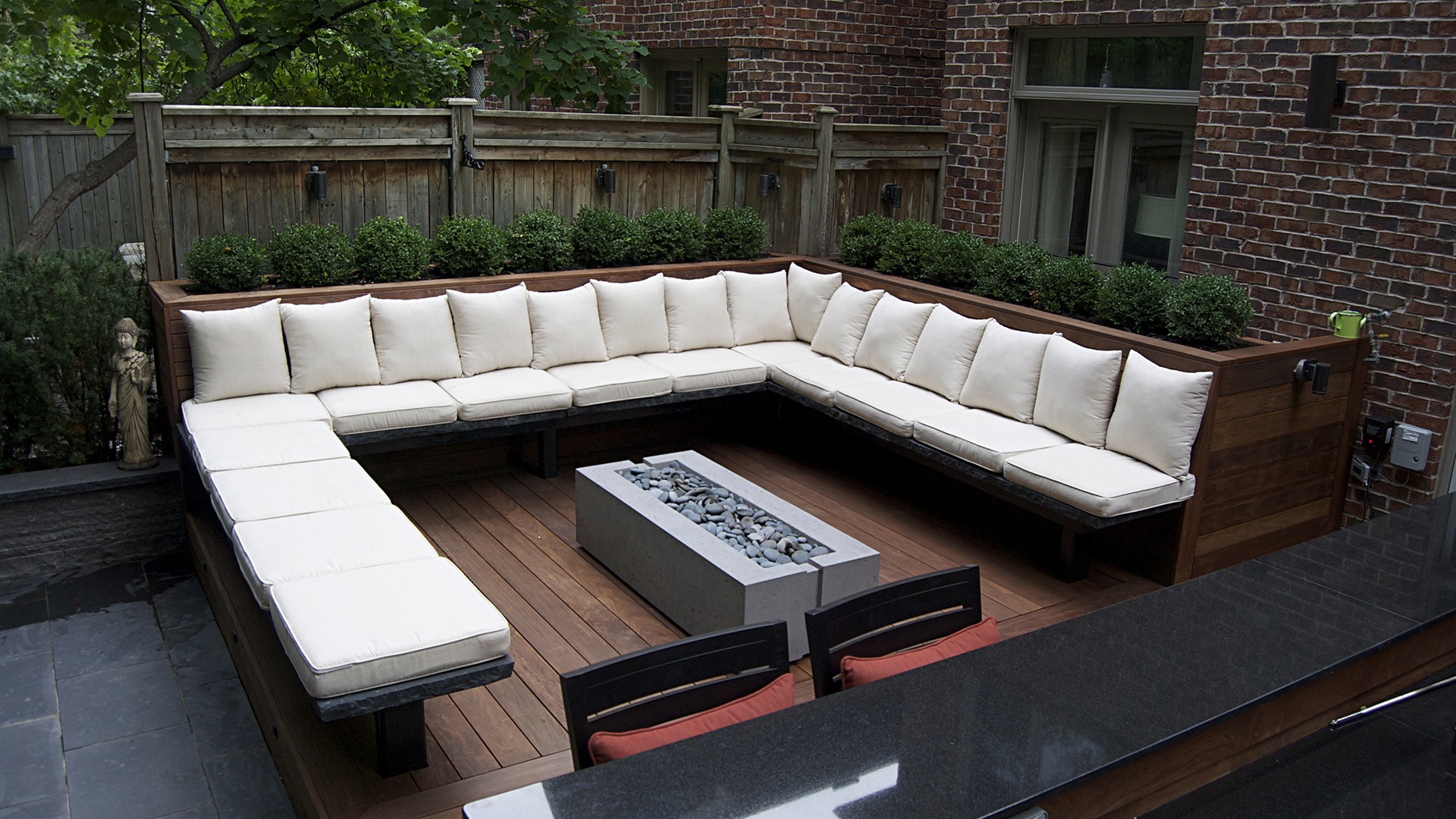 Outdoor patio with an L-shaped cushioned bench, stone fire pit, wooden flooring, surrounded by brick walls and greenery. Quiet, inviting atmosphere.