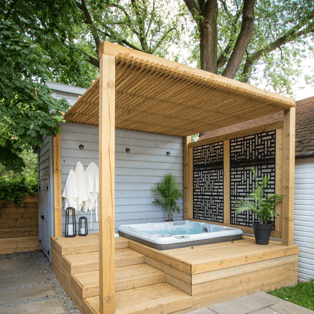 A modern outdoor hot tub under a wooden pergola, surrounded by lush greenery and decorative panels, with towels hanging nearby.