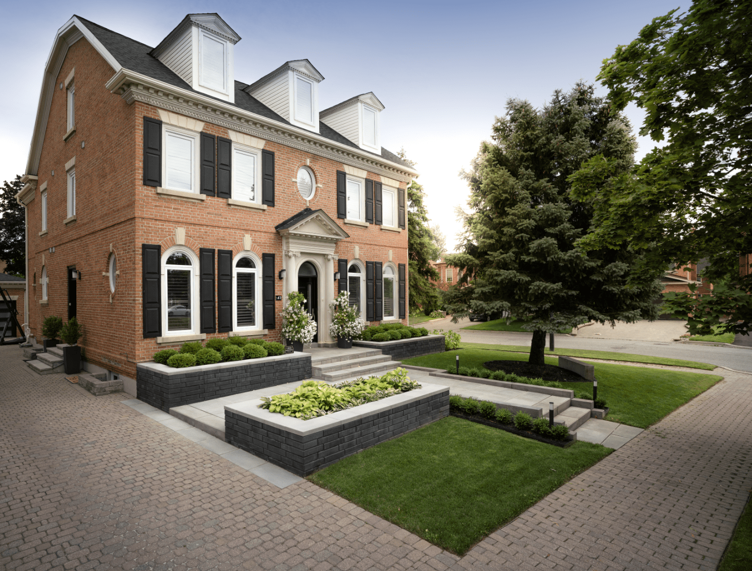 Brick house with black shutters and a manicured lawn situated on a corner lot. Trimmed bushes line the entrance walkway.