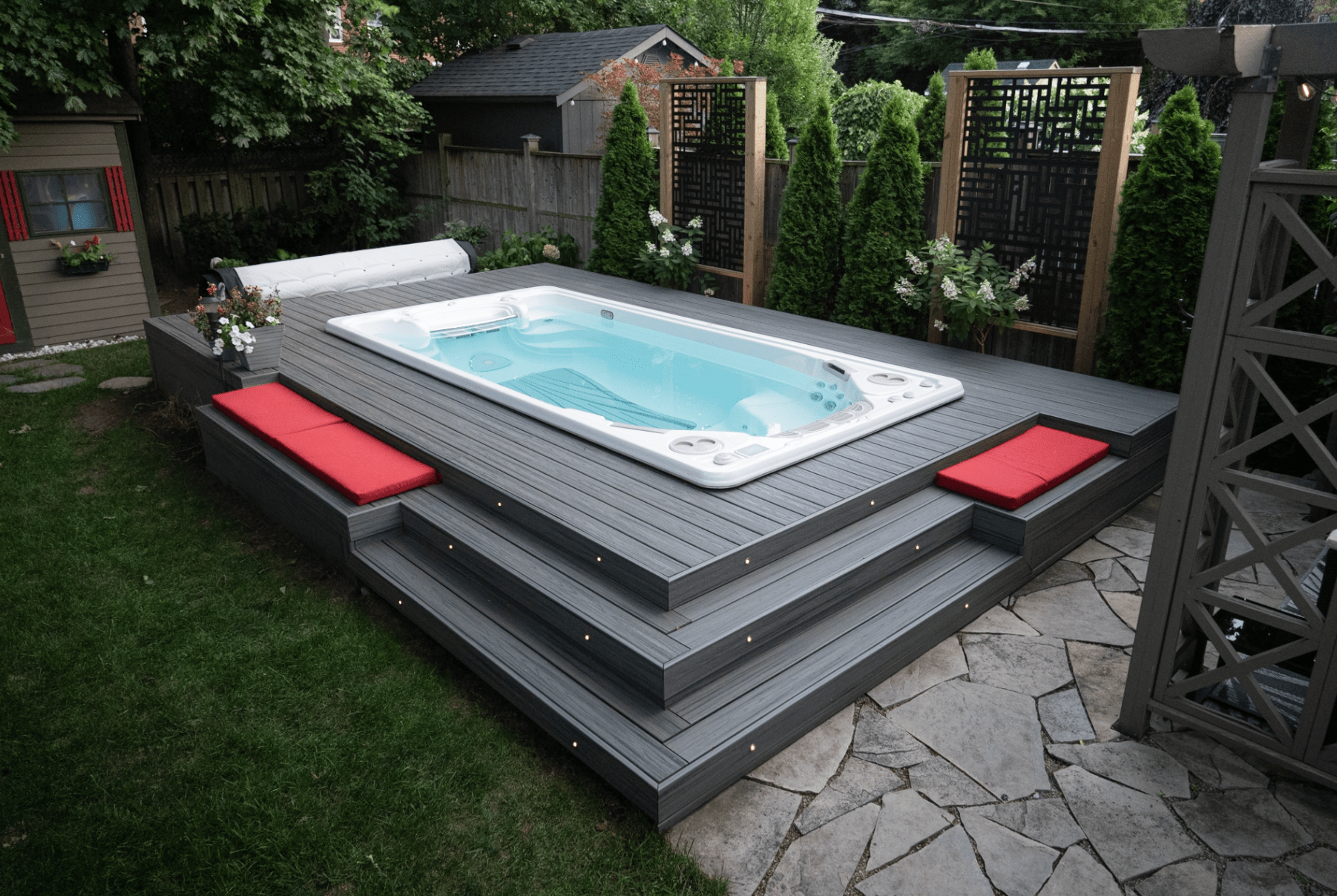 A modern backyard features a raised hot tub with gray decking and red cushions, surrounded by green trees and a small wooden structure.