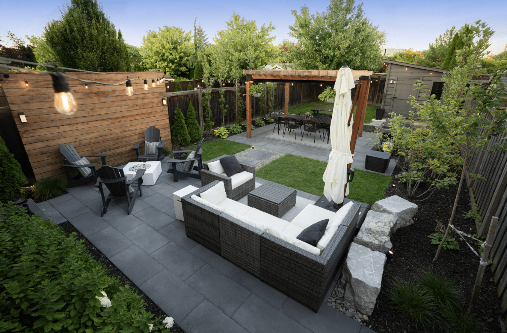 Modern backyard with cozy seating, fire pit, string lights, and lush greenery. Features a wooden pergola, patio furniture, and well-manicured lawn.