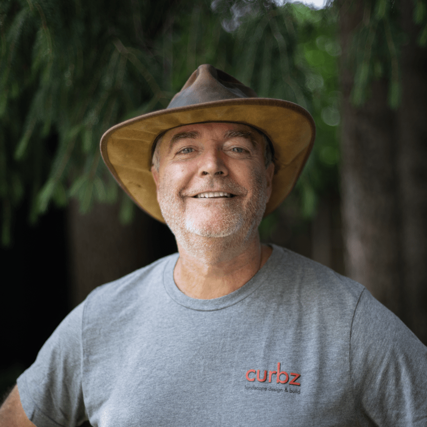 A person wearing a hat smiles outdoors, surrounded by trees, dressed in a gray shirt with "curbz" logo on it.