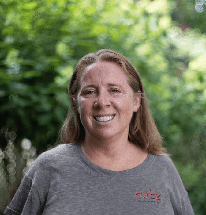 A person smiles outdoors, surrounded by lush green foliage, wearing a gray shirt with a logo. The background features bright, blurred leaves.