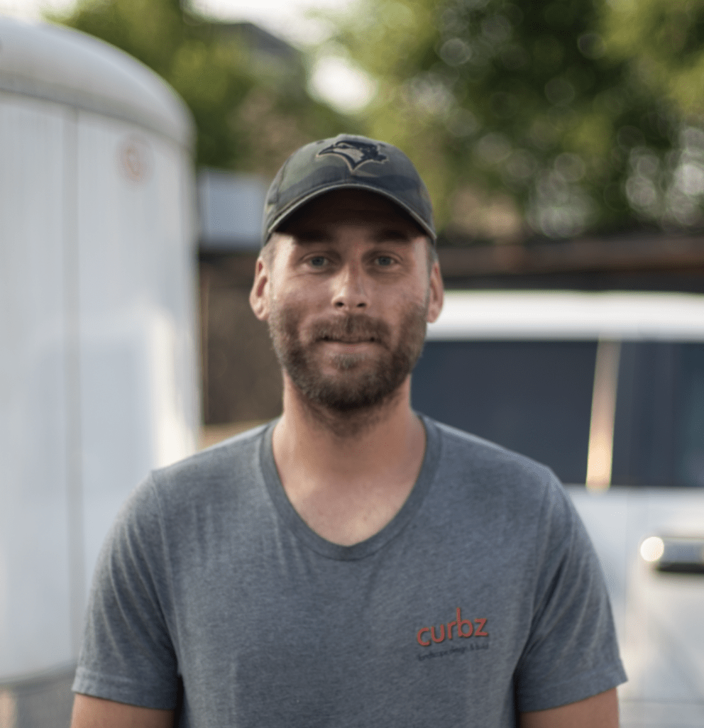A person wearing a cap and gray T-shirt stands outdoors in front of a trailer and vehicle, with trees in the background.