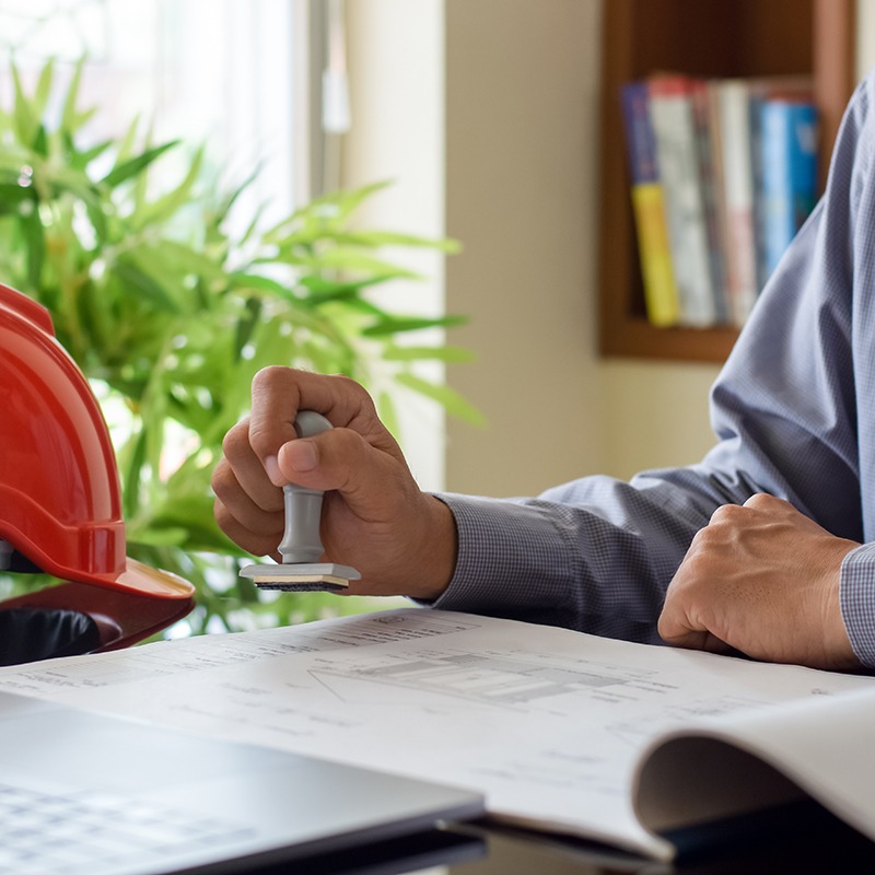 A person stamping architectural blueprints on a desk, with a red hard hat, a laptop, and books in the background.