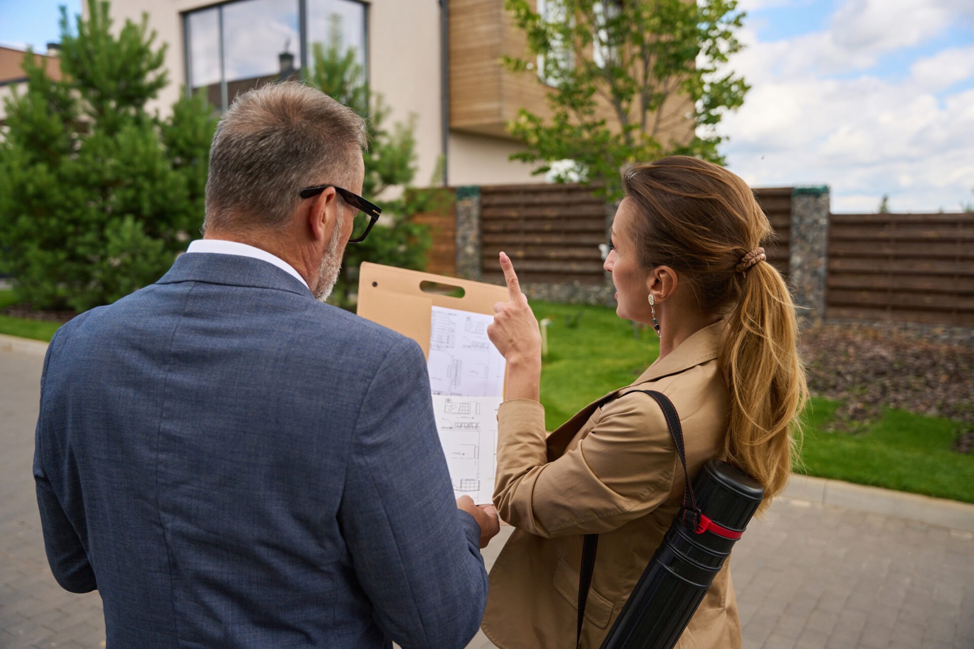 Two people discuss architectural plans outdoors, near a modern building with trees and fencing around. One points, holding a document tube.