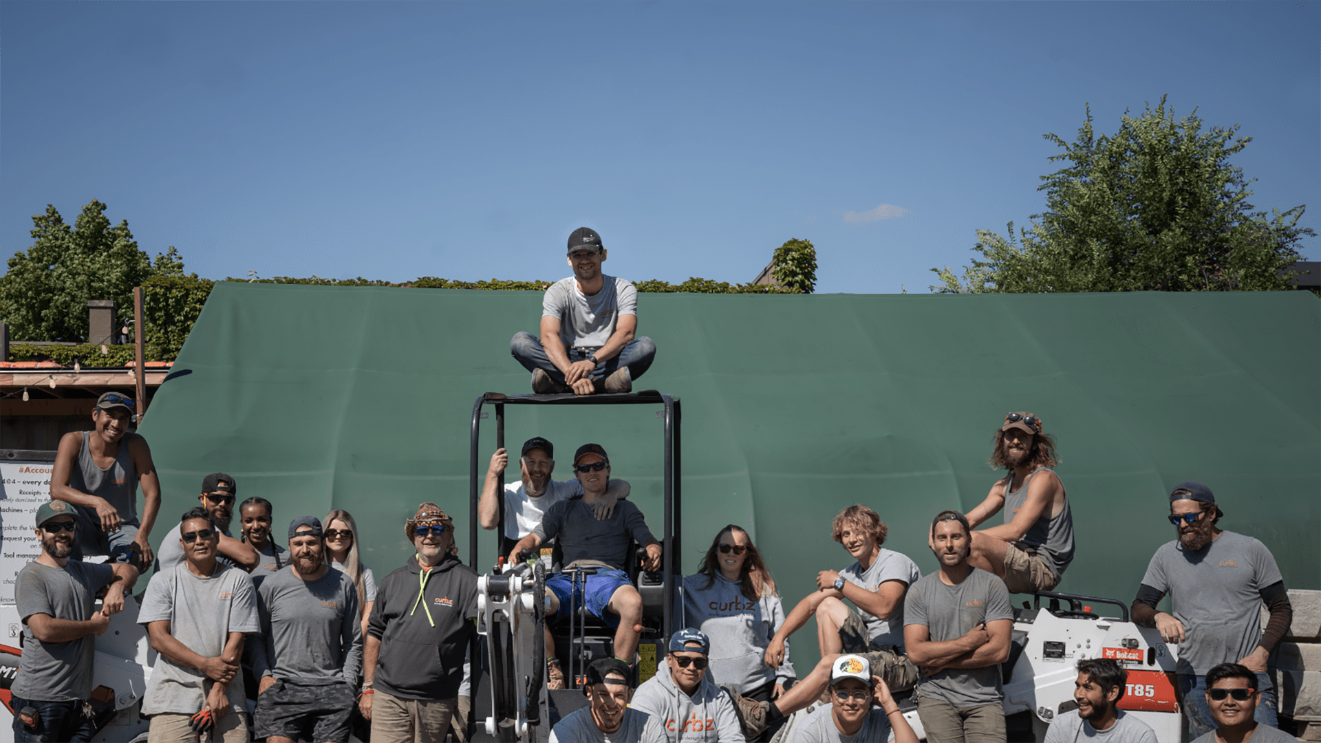 A group of people wearing casual clothing, gathered outdoors around machinery, posing for a photo. Green backdrop and trees visible under blue sky.