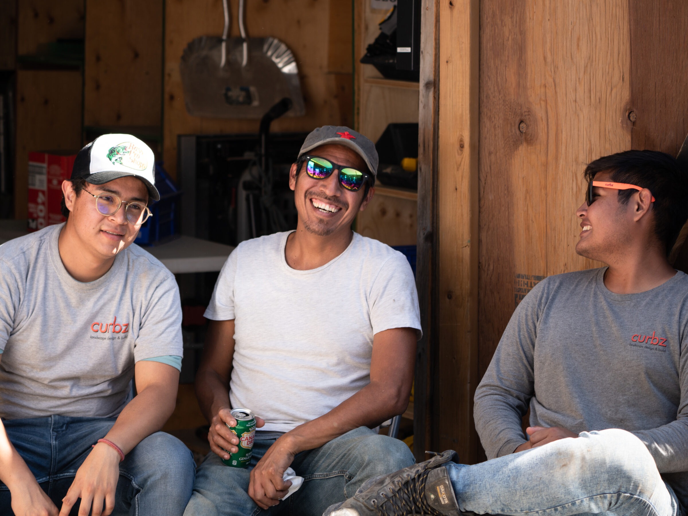 Three smiling people in casual attire sit together outside a wooden structure. One holds a drink can. Relaxed, friendly atmosphere.