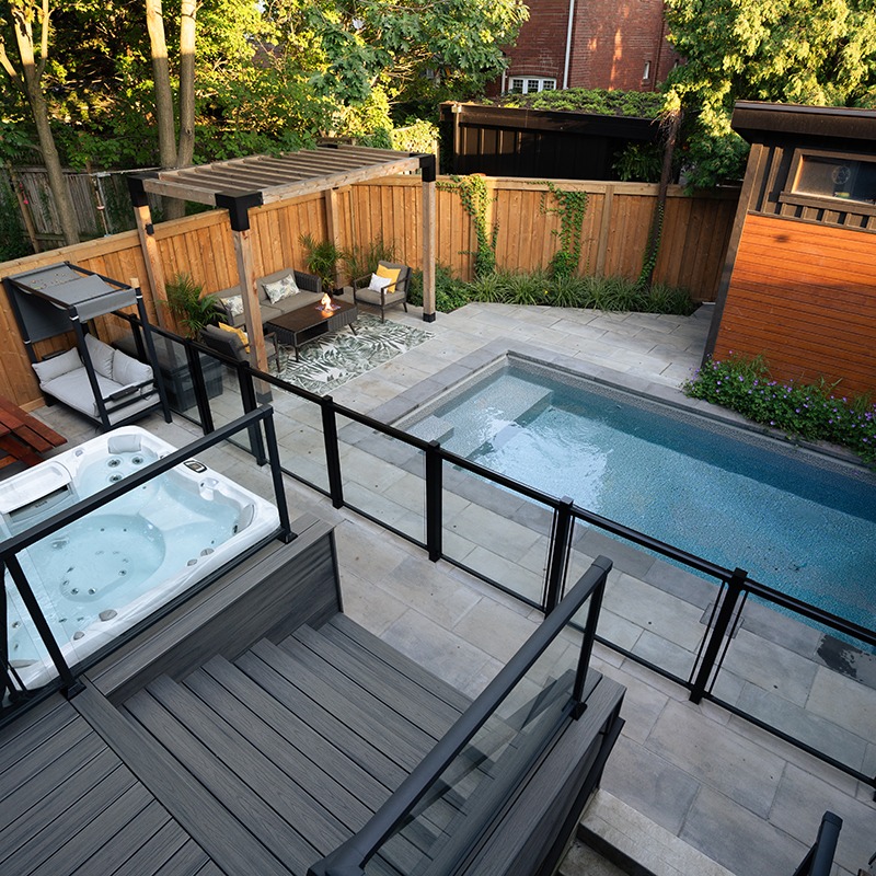A cozy backyard features a pool, jacuzzi, outdoor seating under a pergola, and lush greenery enclosed by a wooden fence.