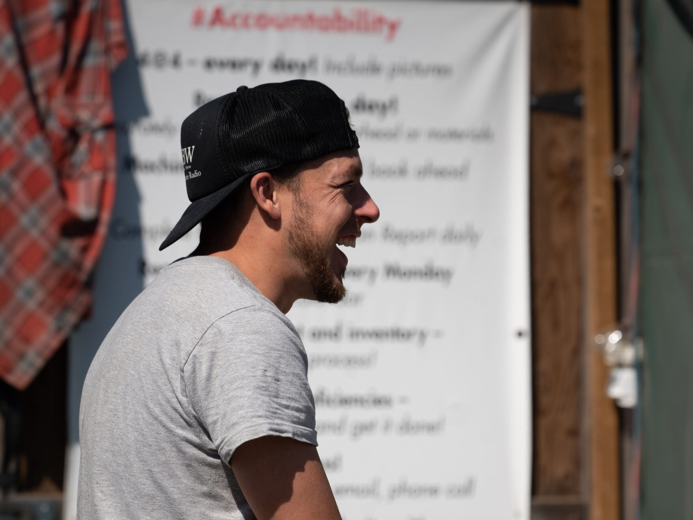 A person in a cap laughs outdoors near a blurred checklist and a red plaid shirt, under sunlight. Background details are indistinct.