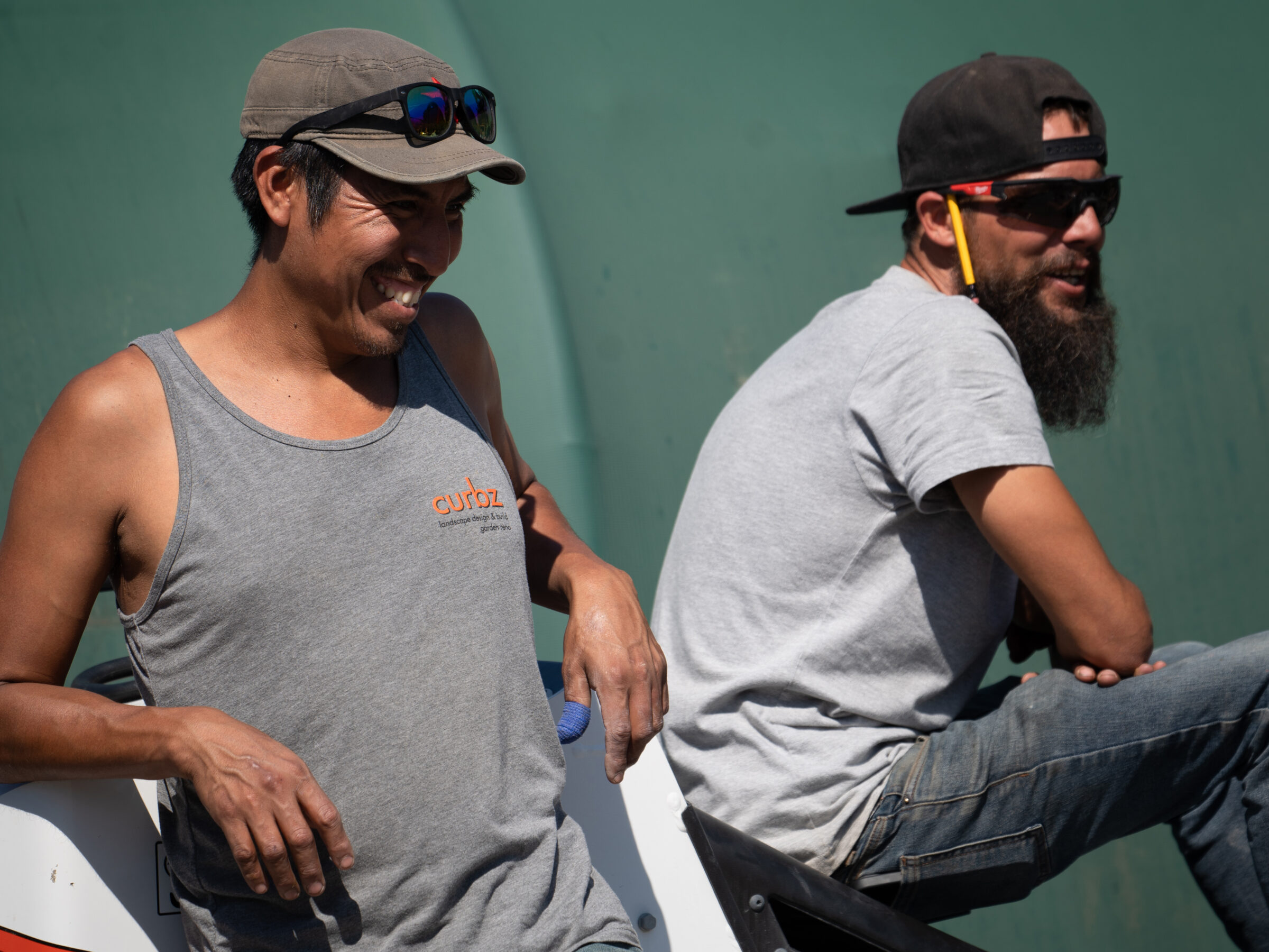 Two people smiling outdoors, wearing casual clothing and sunglasses. They appear relaxed against a green background in a sunny environment. No landmarks visible.