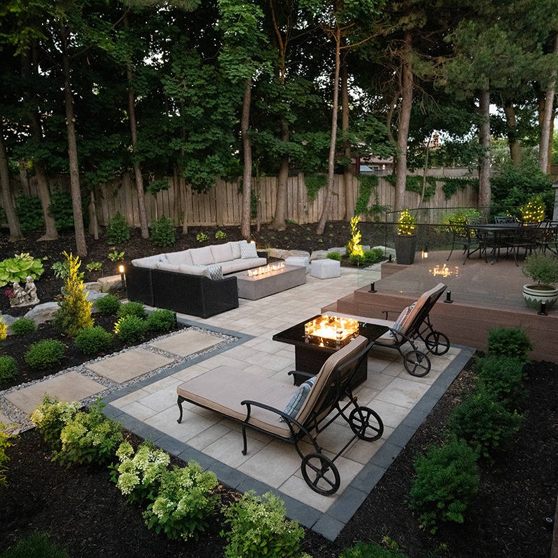 Elegant backyard features of landscaping services in Toronto including cozy seating areas, fire tables, and lush greenery surrounded by tall trees, creating a relaxing outdoor retreat.