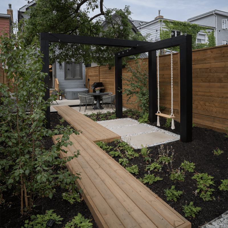 A modern backyard features a wooden pathway, swing, and patio set with chairs, surrounded by plants and an overhead pergola with string lights.