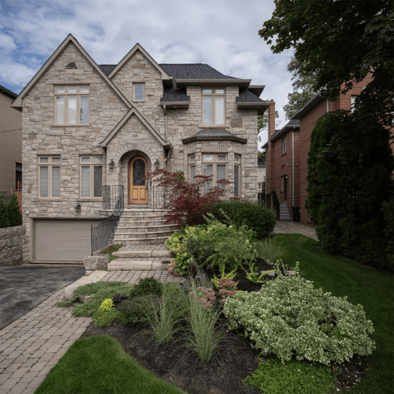 A suburban stone house with a manicured garden and paved driveway, under a cloudy sky, flanked by neighboring red-brick homes.