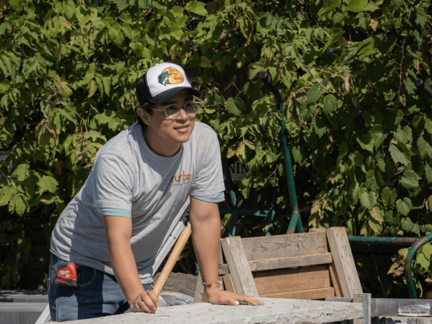 A person in a cap and glasses works outdoors, leaning on a table, surrounded by plants and wooden pallets.