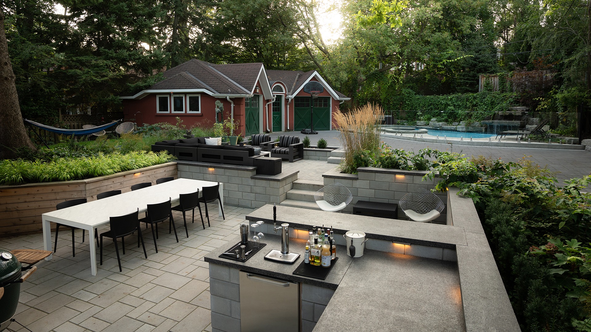 Modern backyard with outdoor kitchen, dining table, and cozy seating area overlooking a large pool surrounded by lush greenery and a red garage.