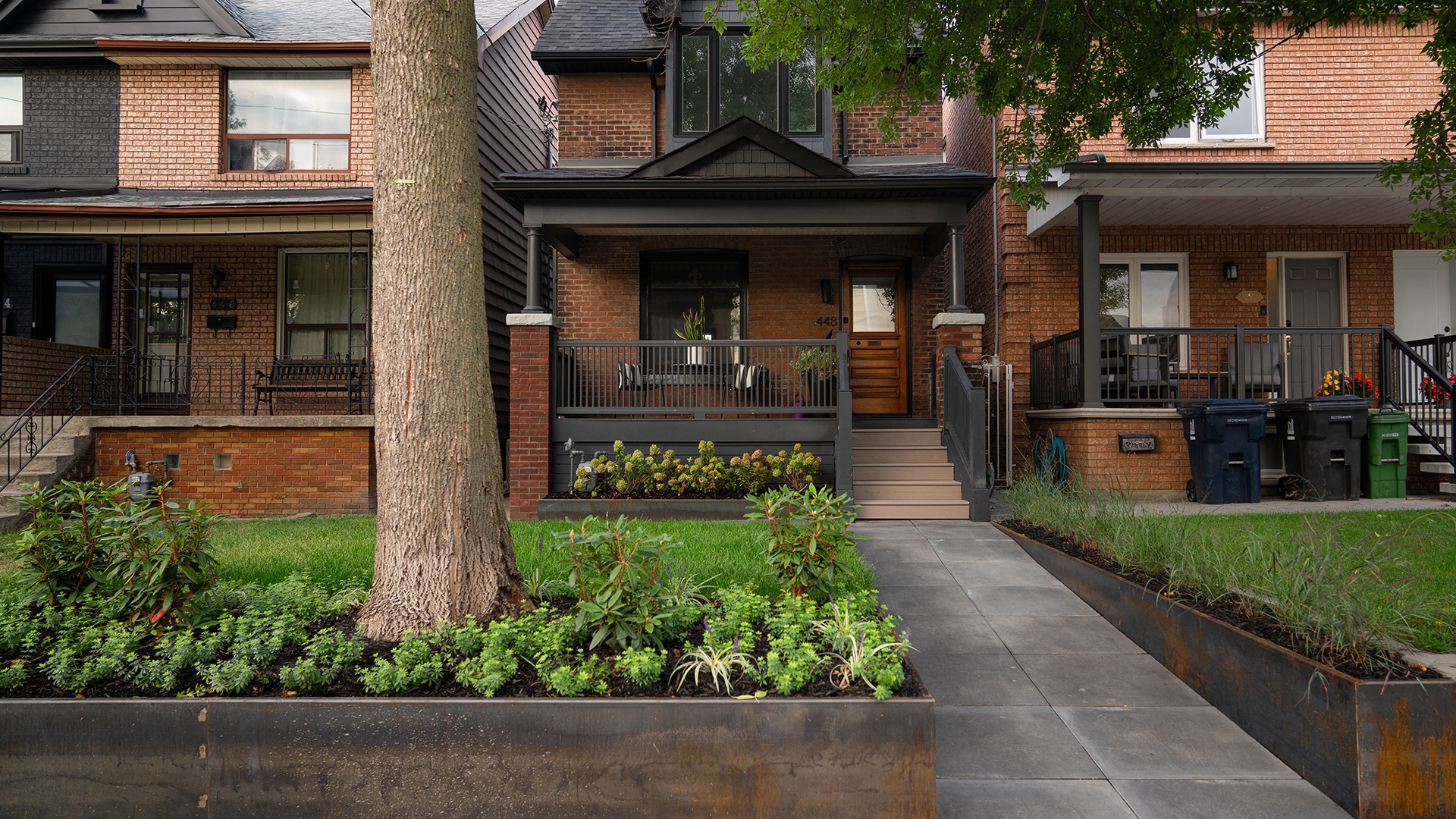 A charming brick townhouse with a cozy porch, surrounded by lush green plants and a large tree in the front yard.