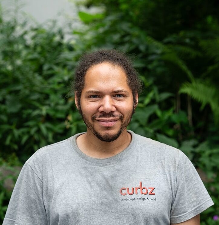 A person with a beard smiles wearing a "curbz" t-shirt, standing in front of lush green foliage. No landmarks or historical buildings visible.