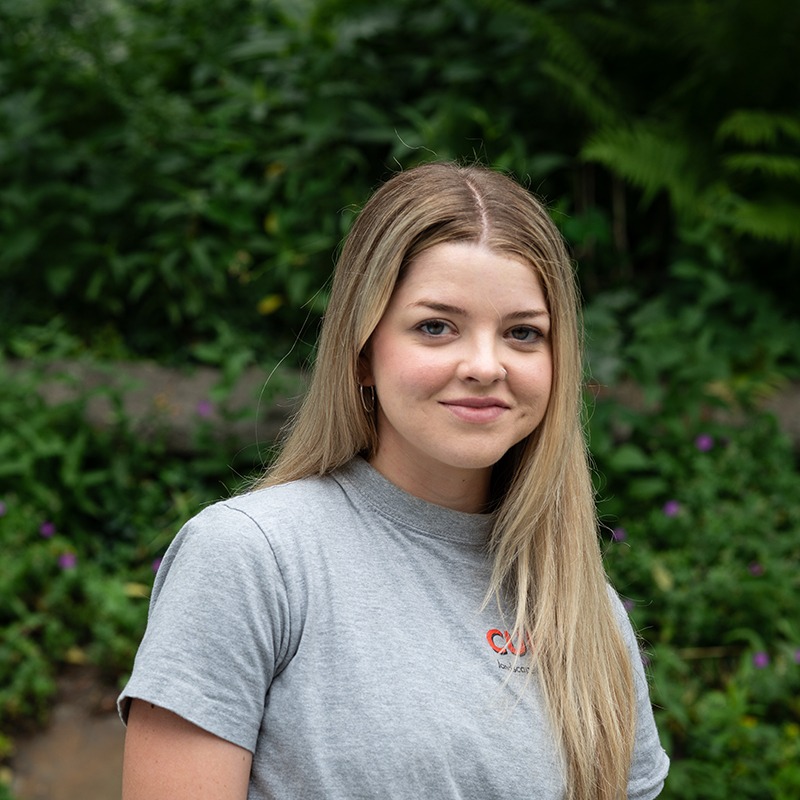 A person with long hair wearing a gray T-shirt stands in front of lush green foliage, looking at the camera.