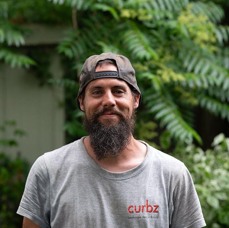 A person with a beard and cap stands in a garden, wearing a gray shirt with "curbz" logo, surrounded by lush greenery.