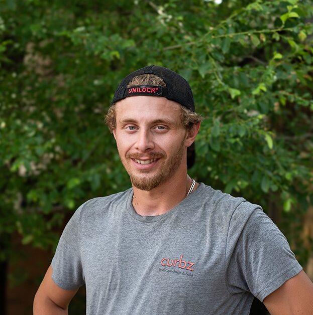 A person wearing a grey shirt and cap stands outdoors, smiling, with a lush green tree background on a sunny day.